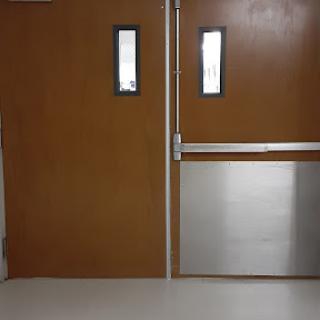 A set of double wooden doors with glass windows, featuring a metal panel, commonly found in a hospital or institutional setting.