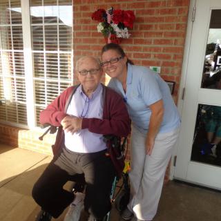 An elderly man in a wheelchair with a caregiver smiling beside him outside a building.