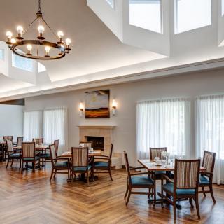 A bright and elegant dining room with multiple tables, a central chandelier, and large windows allowing plenty of natural light.