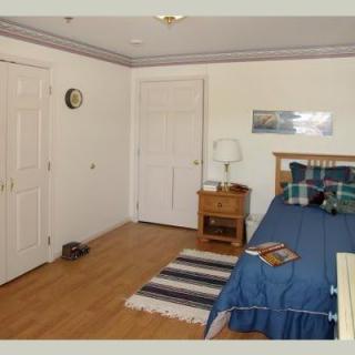 A cozy bedroom with a wooden floor, featuring a neatly made bed with blue bedding, a nightstand with a lamp, and a striped rug.
