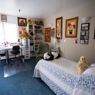A cozy bedroom with artwork on the walls, a bed with plush toys, and a study area by the window.