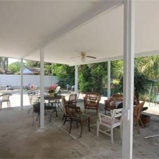 A spacious outdoor patio with various seating arrangements under a covered roof, featuring a ceiling fan and surrounded by greenery.