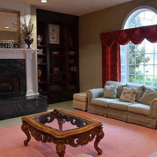 A cozy living room features a plush sofa, an ornate coffee table, and a fireplace adorned with decor. Natural light pours in through a large window framed by red curtains.