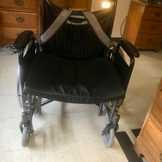 A black wheelchair equipped with a safety harness, positioned on a floor.