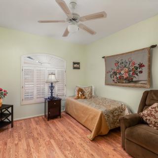 A cozy bedroom with wooden flooring, featuring a single bed, a ceiling fan, and decorative elements like a floral tapestry and window shutters.