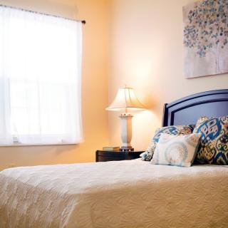 A cozy bedroom with a neatly made bed, stylish cushions, a bedside lamp, and soft natural light coming through the window.