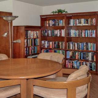 A cozy library room featuring wooden bookshelves filled with books, a round wooden table, and cushioned chairs.