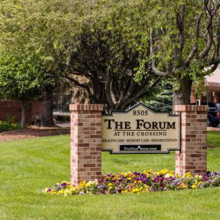 A sign for 'The Forum at the Crossing' surrounded by flowers and trees, marking the entrance to a building complex.