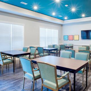 A modern meeting room with multiple tables and chairs, featuring bright lighting and a television on the wall.