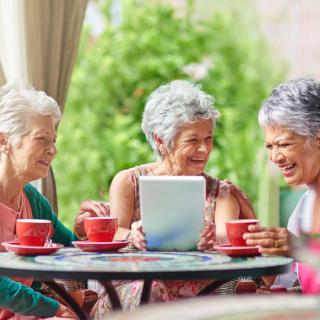 Three elderly women enjoying coffee and conversation while using a tablet outdoors.