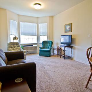 A cozy living room with a couch, armchairs, and a television, illuminated by natural light from large windows.