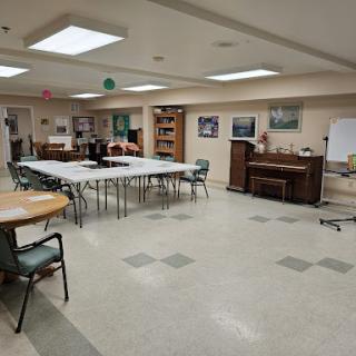 A spacious community room set up with tables and chairs, featuring a piano and artwork on the walls.