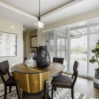A modern dining room with a round table and stylish decor, featuring large windows with blinds and an indoor plant.