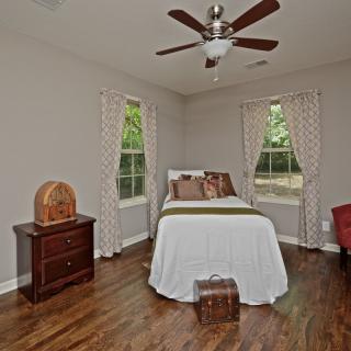 A cozy bedroom with a wooden floor, featuring a bed, two windows with patterned curtains, a bedside table, and a ceiling fan.
