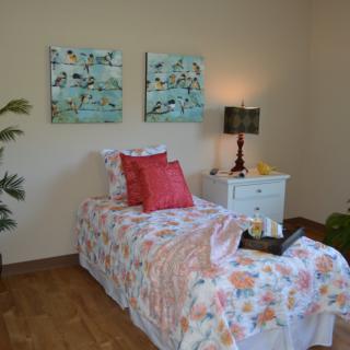A cozy bedroom with a single bed featuring colorful bedding and decorative pillows, accompanied by a nightstand with a lamp and vibrant artwork on the walls.