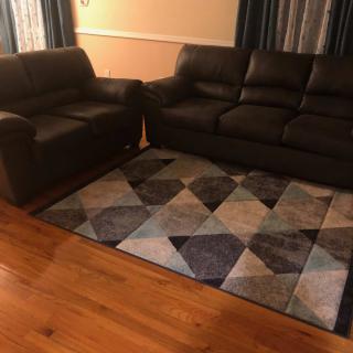 A cozy living room setup featuring a dark couch, loveseat, and a geometric patterned rug on hardwood flooring.