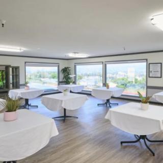 A bright dining room with round tables covered in white tablecloths, large windows, and decorative plants.