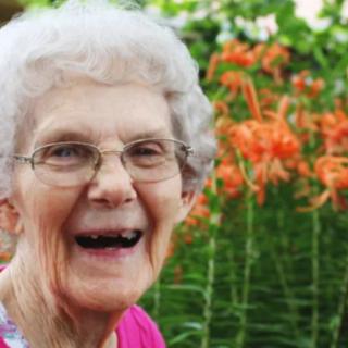 An elderly woman smiling in a garden with orange flowers in the background.