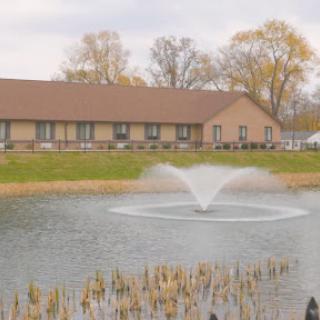 A serene view of a pond featuring a decorative fountain, surrounded by autumn foliage and a modest building.