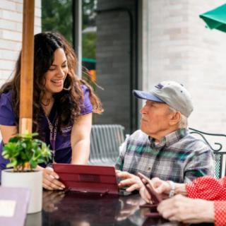 An outdoor scene where a woman is engaging in conversation with an elderly man and woman, assisting them with a tablet.