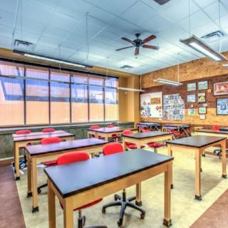 An empty classroom with desks and chairs neatly arranged, featuring large windows and ceiling fans.