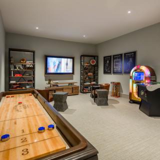 A comfortable game room featuring a shuffleboard table, vintage arcade machine, jukebox, and television with a cozy seating area.