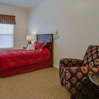 A cozy bedroom with a red bedspread, patterned armchair, and lamp by the window.