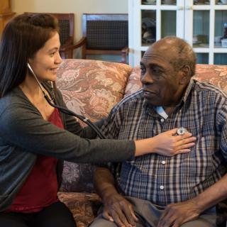 A nurse uses a stethoscope to check the heart of an elderly man during a home healthcare visit.