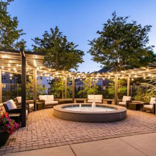 A cozy outdoor patio with a central fire pit, surrounded by comfortable seating and illuminated by string lights, set against an evening backdrop of trees.