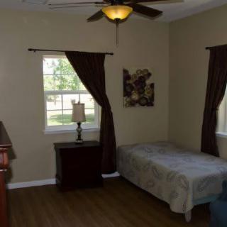 A cozy bedroom featuring a single bed, nightstand with lamp, and windows with dark curtains.