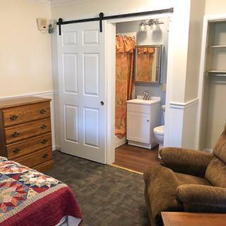 A cozy bedroom with a barn door leading to an adjacent bathroom, featuring a bed, wooden dresser, and armchair.