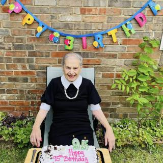 An elderly woman celebrating her birthday outdoors with a cake and decorations.