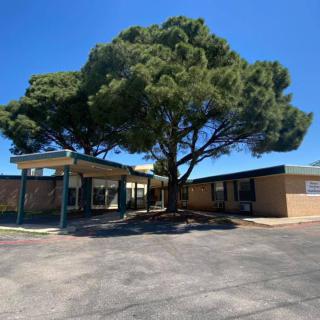 A school building with a large tree in the foreground under a clear blue sky.
