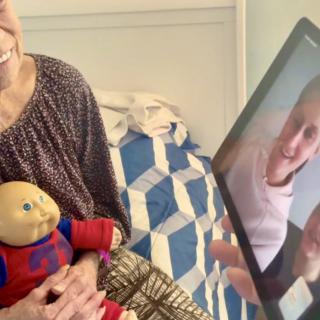 A person holding a doll is on a video call with two people displayed on a laptop screen in a bedroom setting.
