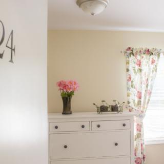 A cozy bedroom with a white dresser, floral curtains, and a vase of pink flowers.