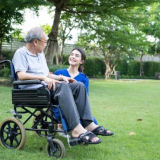An elderly man in a wheelchair enjoys a conversation with his caretaker in a park setting.