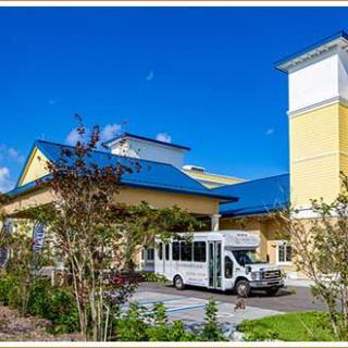 A yellow building with a blue roof and a white bus parked in front, surrounded by trees under a clear blue sky.
