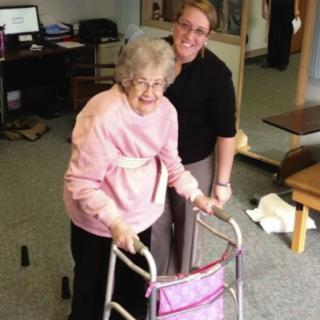 An elderly woman using a walker, accompanied by a caregiver indoors.
