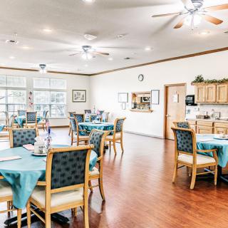A spacious and well-lit dining area with neatly arranged tables and chairs, featuring a kitchen area in the background.