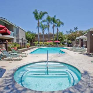 A sunny outdoor residential area featuring a swimming pool and hot tub, surrounded by palm trees and apartment buildings.