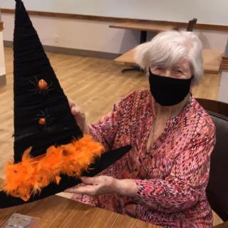 An elderly woman wearing a mask holds a festive witch hat adorned with orange feathers, celebrating Halloween indoors.