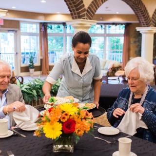 A waitress serves an elderly couple in a restaurant setting, with a colorful floral arrangement on the table.