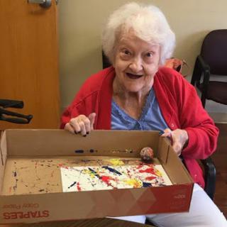 An elderly woman joyfully displays her artwork after an engaging painting session.