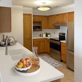 A modern kitchen with wooden cabinets, stainless steel appliances, and a clean white countertop featuring a fruit bowl.