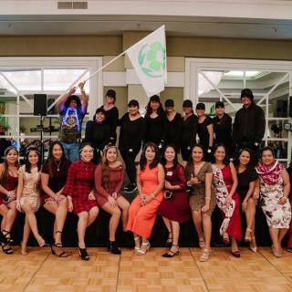 A group of people dressed in formal attire pose together indoors, some holding a flag.