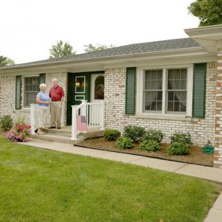 A classic brick house with a well-maintained garden and two people standing on the porch.