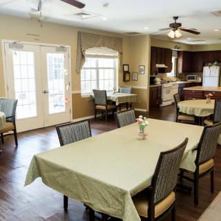 A cozy dining area with multiple tables and chairs, connected to a kitchen space, featuring large windows and ceiling fans.