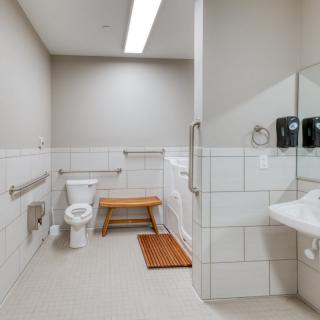 A clean and modern accessible bathroom with a toilet, sink, handrails, and a wooden bench.