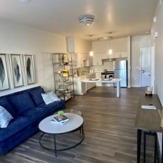 A modern living room with an open floor plan featuring a blue sofa, wood flooring, and a kitchen area.