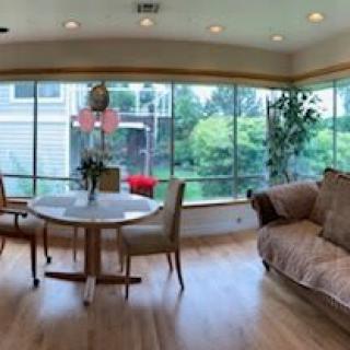 A bright dining and living area with large windows, allowing natural light to fill the space. The room features a round table with chairs, a comfortable sofa, and indoor plants on a hardwood floor.